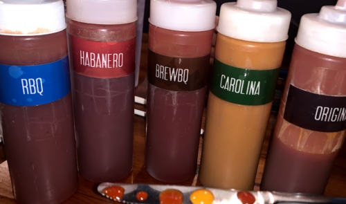Five squeeze bottles of different sauces labeled RBQ, Habanero, BrewBQ, Carolina, and Original, on a wooden table.