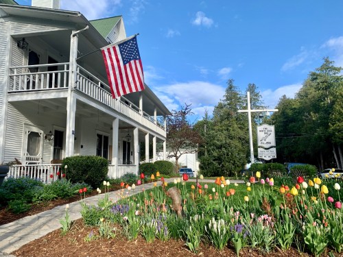 A white house with a porch, an American flag, and a colorful flower garden in front under a blue sky.