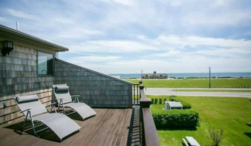 A balcony with two lounge chairs overlooks a grassy area and the ocean under a clear blue sky.