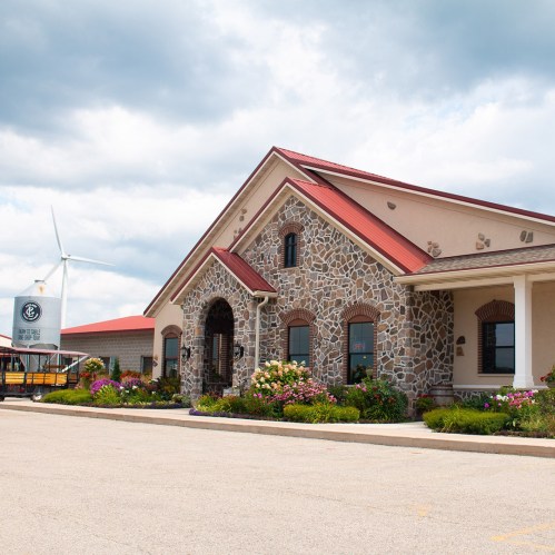A stone building with a red roof, surrounded by flowers, with a wind turbine in the background under a cloudy sky.