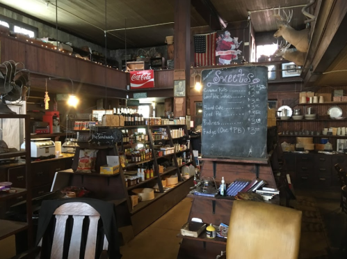 Interior of a rustic store with wooden shelves, a chalkboard menu, and various products displayed. Cozy, vintage atmosphere.