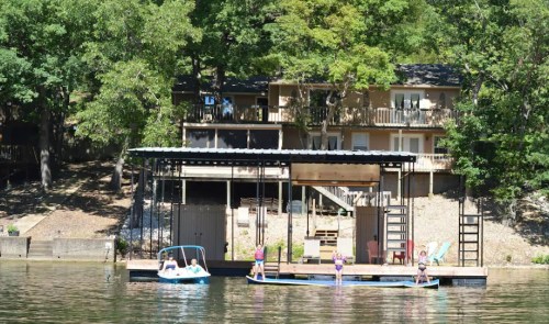 A lakeside scene featuring a dock, a boat, and people enjoying the water, with a house surrounded by trees in the background.