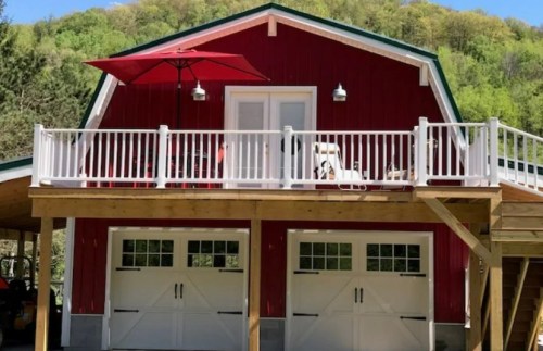 A red barn-style house with a deck, white railings, and an umbrella, surrounded by green trees.