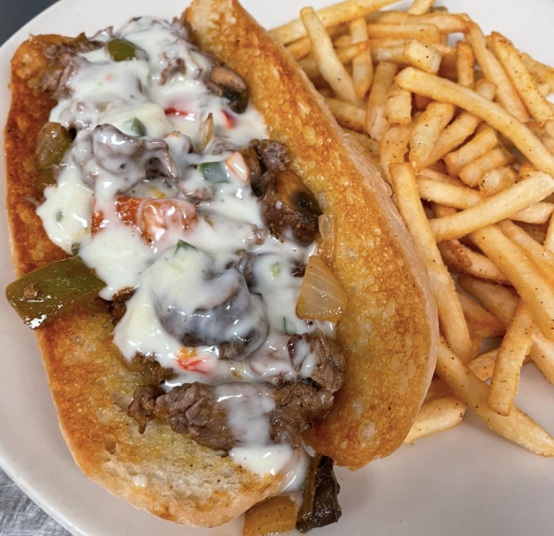 A loaded cheesesteak sandwich with sautéed vegetables and a side of crispy French fries.