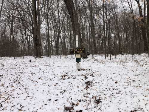 A snowy forest scene with a signpost for the Little Falls Trail amidst bare trees and a white ground cover.