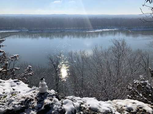 A snowy landscape overlooking a river, with a small snowman in the foreground and trees lining the opposite bank.