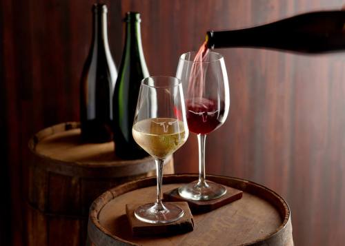 Two wine glasses on a wooden barrel, one filled with white wine and the other with red, with bottles in the background.