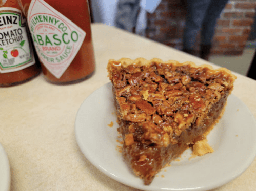 A slice of pecan pie on a plate, with bottles of ketchup and hot sauce in the background.