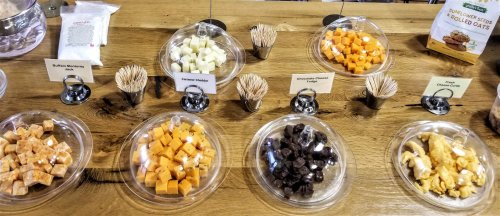 A display of assorted cheeses and snacks in clear bowls on a wooden table, labeled with names and accompanied by toothpicks.