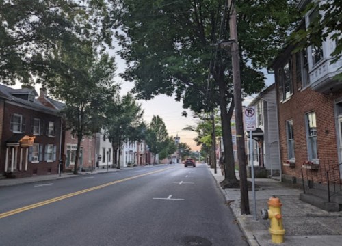 Quiet street lined with trees and houses, featuring a clear sky and a parked car near a fire hydrant.