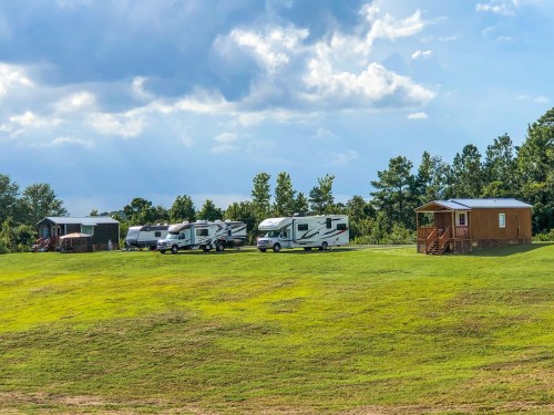 A grassy area with several RVs parked and a small cabin in the background under a partly cloudy sky.