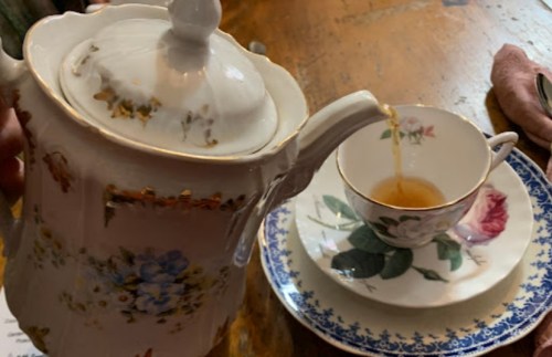 A teapot pours tea into a delicate floral teacup on a patterned saucer, set on a wooden table.