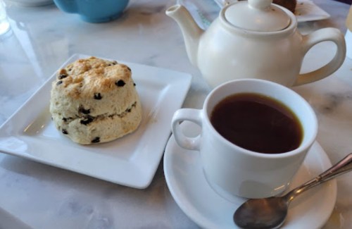A cup of tea beside a scone on a white plate, with a teapot in the background on a marble table.