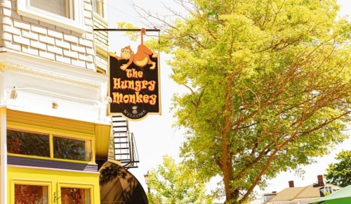 Sign for "The Hungry Monkeys" hanging from a building, surrounded by green trees on a sunny day.