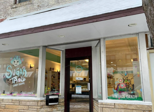 Exterior of Sugar Troll Candy Shoppe, featuring large windows and a sign, showcasing vintage candy and a cozy atmosphere.