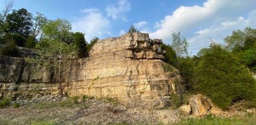 A rocky cliff surrounded by greenery under a partly cloudy sky.