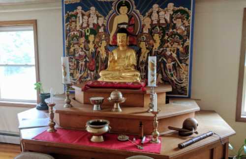 A golden Buddha statue on a wooden altar, surrounded by candles and traditional decor, with a colorful backdrop.