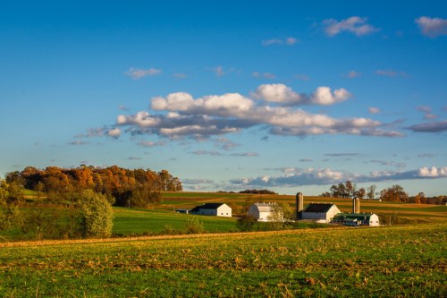 A picturesque farm landscape with green fields, a few buildings, and colorful autumn trees under a blue sky with clouds.