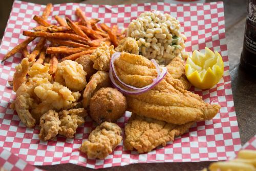 A platter of fried seafood, including fish, shrimp, and hushpuppies, served with sweet potato fries and a lemon wedge.