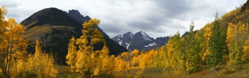 A panoramic view of mountains with snow, surrounded by vibrant autumn trees in shades of yellow and green under a cloudy sky.