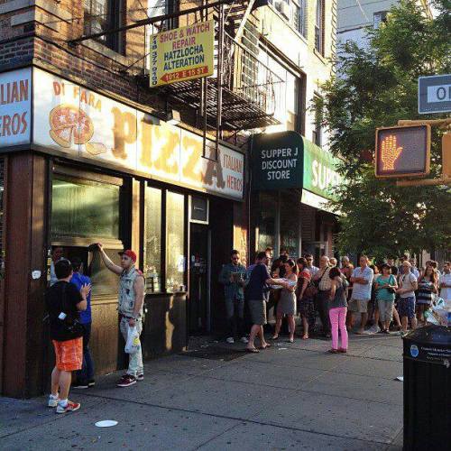 A busy street scene with a long line outside a popular pizzeria and a discount store nearby.