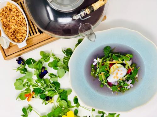 A colorful salad with greens, edible flowers, and a poached egg, accompanied by a teapot and granola on a wooden tray.