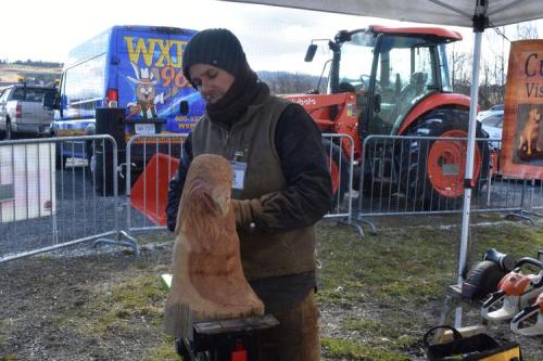 A man carves a wooden sculpture of a lion's head at an outdoor event, with a tractor and vehicles in the background.