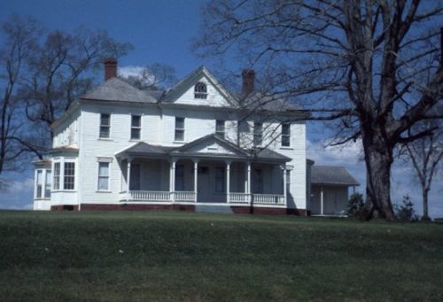 A large, white historic house with a porch, set against a blue sky and green lawn, surrounded by trees.
