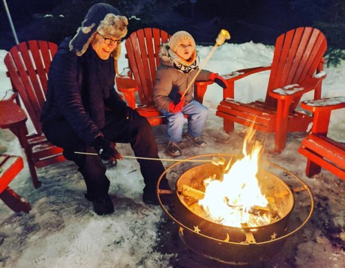 A person and a child sit by a fire pit, roasting marshmallows, surrounded by snow and red Adirondack chairs.