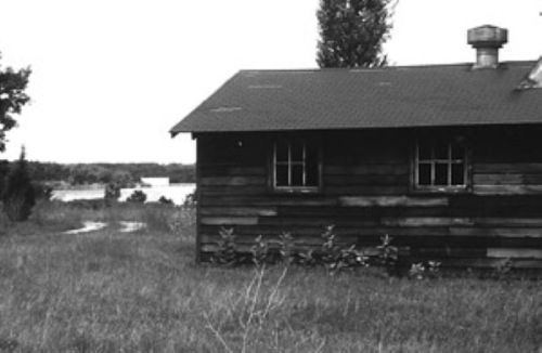 A weathered wooden cabin beside a grassy area, with a view of a lake in the background under a cloudy sky.