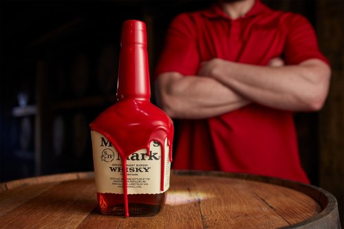A bottle of whiskey with red wax dripping down, placed on a wooden barrel, with a person in a red shirt in the background.