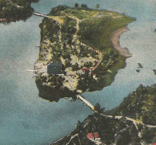 Aerial view of a small island with a building, surrounded by water and greenery, featuring a bridge connecting to the mainland.