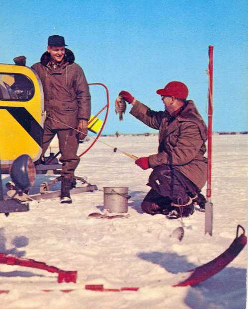 Two men ice fishing on a snowy landscape, one holding a fish while the other smiles near a snowmobile.