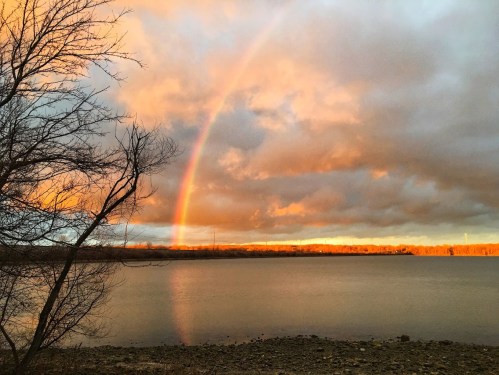 A serene landscape featuring a rainbow arching over a calm lake at sunset, with trees silhouetted in the foreground.