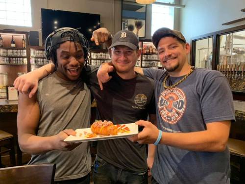 Three men smiling and posing together in a restaurant, holding a plate of sushi.