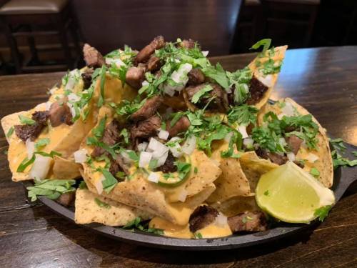 A plate of nachos topped with grilled meat, cheese, onions, cilantro, and lime wedges on a wooden table.