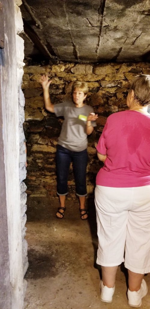 A woman gestures while speaking in a dimly lit stone room, with another woman listening attentively.