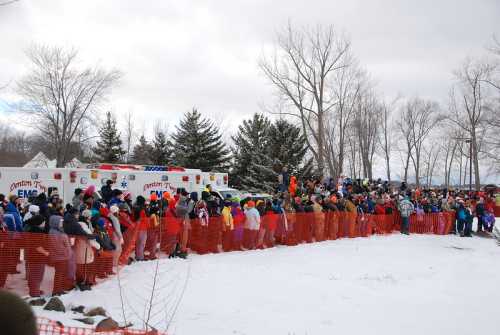 A large crowd gathers behind a snow fence, with emergency vehicles in the background and trees lining the scene.