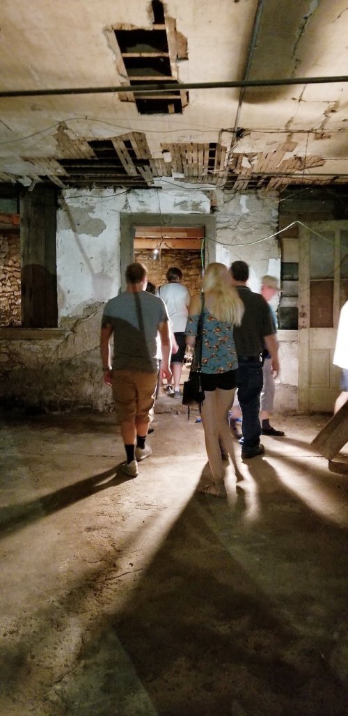A group of people walking through a dimly lit, old basement with exposed walls and a doorway ahead.