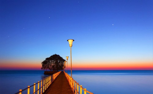 A serene pier extends into calm waters under a twilight sky, with stars twinkling above and a tree at the end.