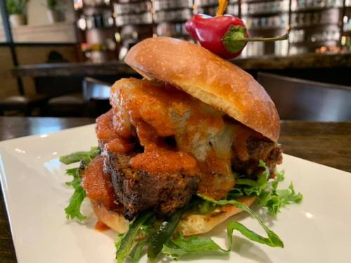 A hearty sandwich with layers of meat, sauce, and greens, topped with a cherry pepper on a toasted bun.