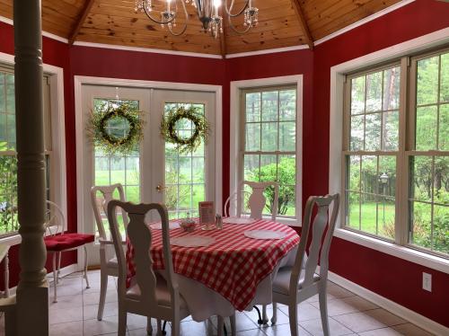 Cozy dining area with a checkered tablecloth, white chairs, and large windows overlooking a green garden.