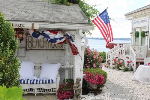 Charming seaside cottage with an American flag, flowers, and a cozy seating area by the water.