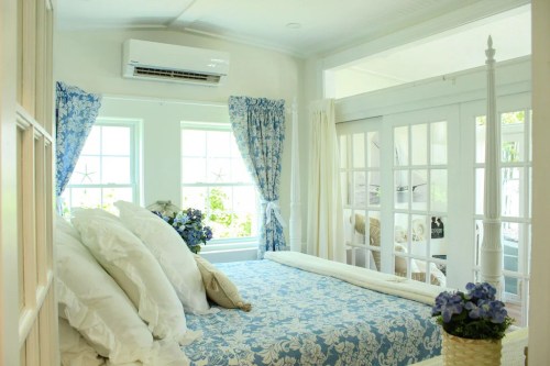 Bright bedroom with floral bedding, large windows, and blue curtains, featuring a cozy atmosphere and decorative flowers.