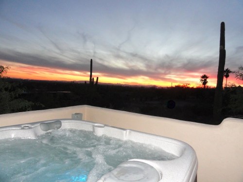 A hot tub with bubbling water overlooks a vibrant sunset and desert landscape with cacti and palm trees.