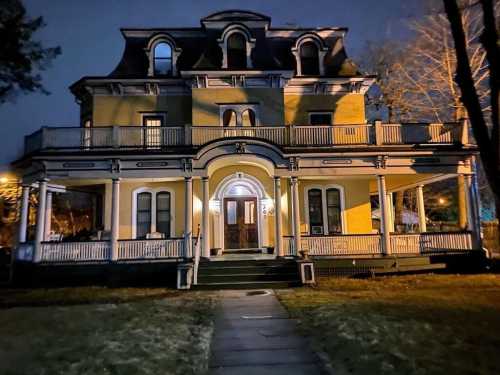 A large, historic house with a wraparound porch, illuminated at night, surrounded by trees and a grassy lawn.