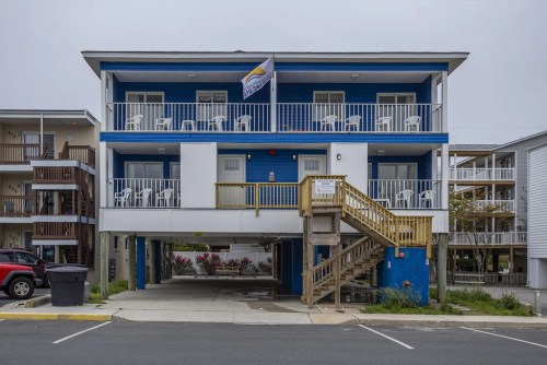 A blue and white two-story building with balconies, featuring white chairs and a flag, located near a parking area.