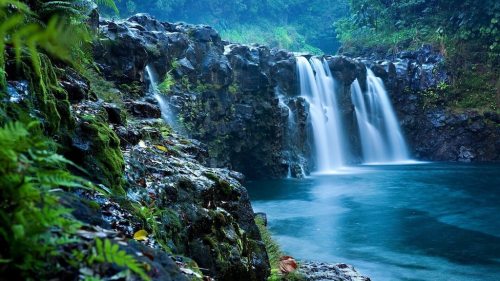 A serene waterfall cascades over rocky cliffs into a tranquil blue pool, surrounded by lush greenery.