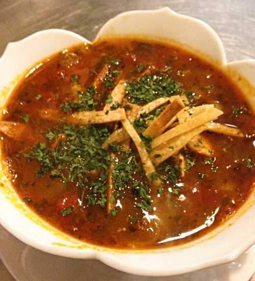 A bowl of soup garnished with tortilla strips and fresh herbs, featuring a rich, colorful broth.