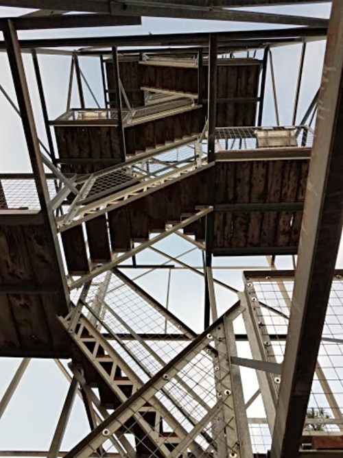 A spiral staircase viewed from below, featuring metal and wooden steps with a clear sky above.